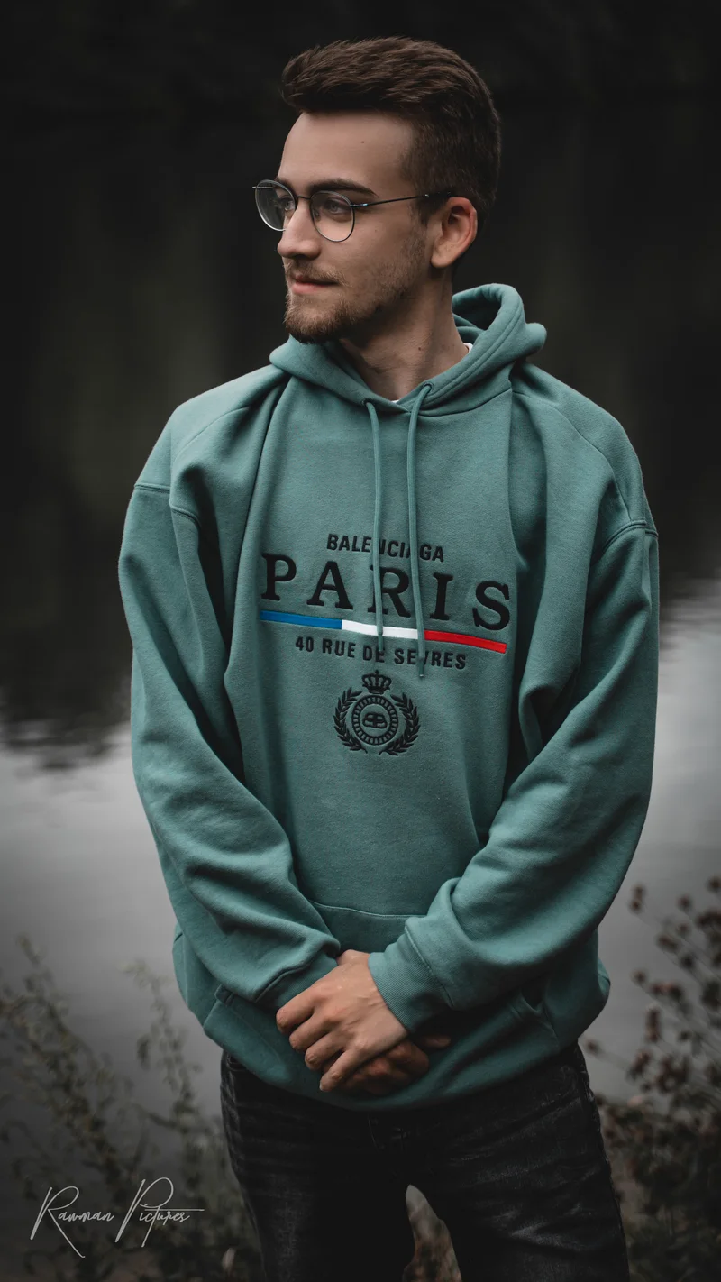 RawmanPictures - Sebastian Schiefermayr | Shooting Philipp B. | Porträtfotografie im Freien: Stilvoller junger Mann am Wasser mit Kapuzenpullover und Brille.