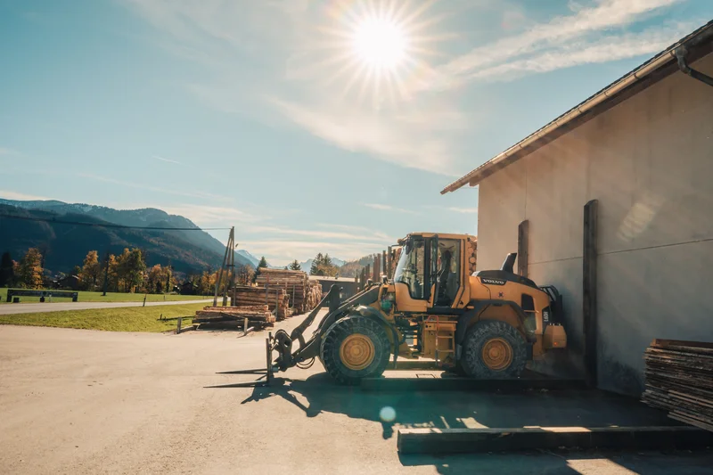 RawmanPictures - Sebastian Schiefermayr | Sägewerk Löberbauer | Sonnenbeschienene Landschaft mit Baugerät und Bergen im Hintergrund. Ideal für Industrie- und Landschaftsfotografie.