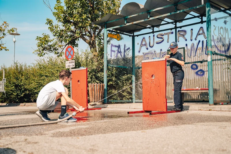 RawmanPictures - Sebastian Schiefermayr | Sommereisdisco | Zwei Personen üben mit Wasserschläuchen an einer Bushaltestelle unter blauem Himmel.