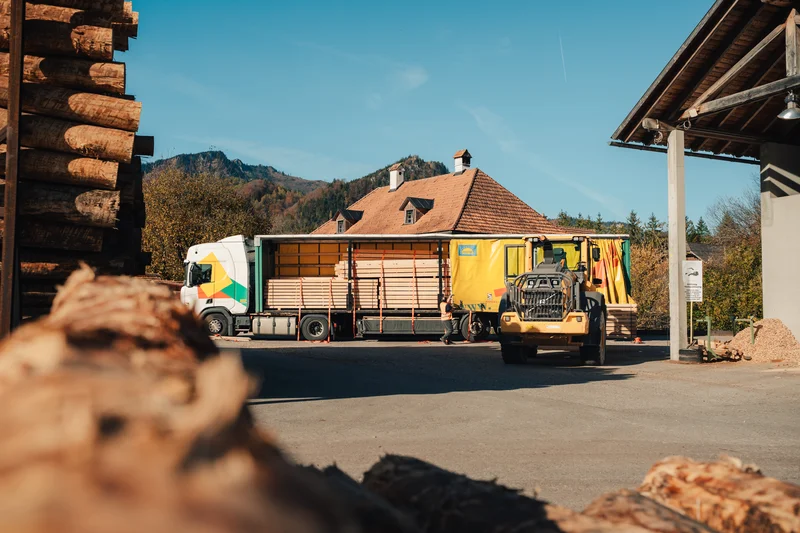 RawmanPictures - Sebastian Schiefermayr | Sägewerk Löberbauer | Transport von Holzstämmen im Sägewerk, eingebettet in malerische Bergkulisse.
