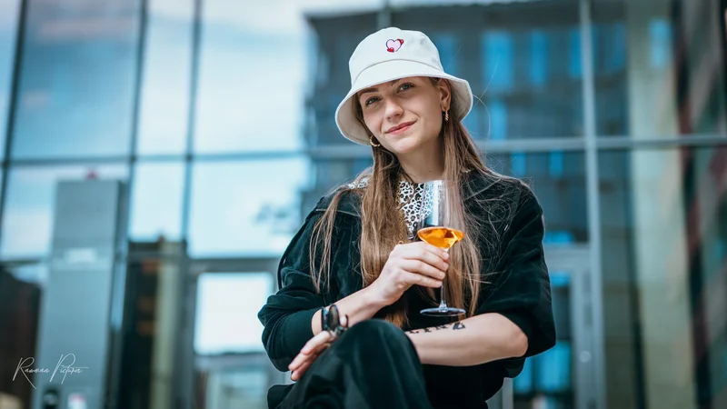 RawmanPictures - Sebastian Schiefermayr | Musikvideodreh "Flinz Asozial" | Moderne Porträtfotografie: Lächelnde Frau mit Hut und Drink im urbanen Hintergrund.