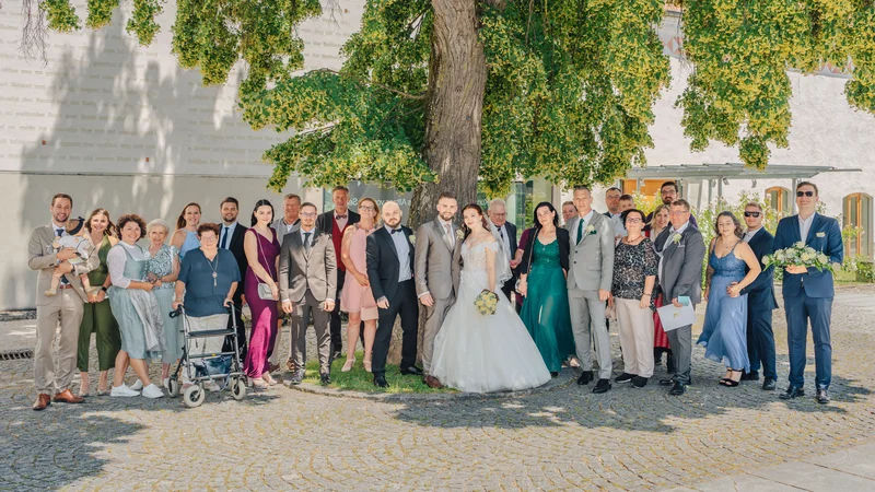 RawmanPictures - Sebastian Schiefermayr | Hochzeit Natalia & Jürgen | Hochzeitsgesellschaft im Freien vor Baum, festliche Kleidung, sonniger Tag, Gemeinschaft.