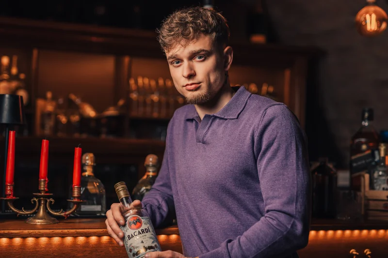 RawmanPictures - Sebastian Schiefermayr | Portraitshooting Kevin F. | Junger Mann in Bar, hält Bacardi-Flasche im warmen Licht. Lifestyle-Fotografie, Bar-Kultur.