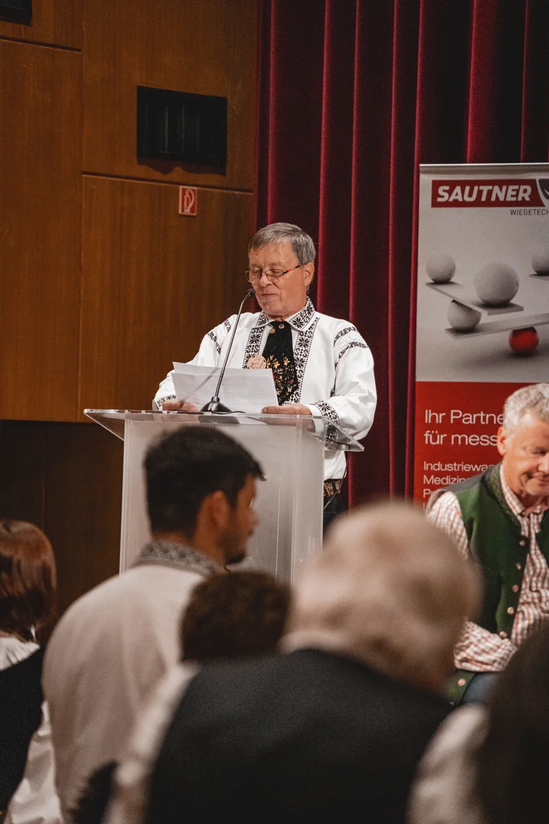 RawmanPictures - Sebastian Schiefermayr | Volkstanzfest | Festliche Veranstaltung mit traditioneller Kleidung, Moderation am Rednerpult, kulturelles Event.