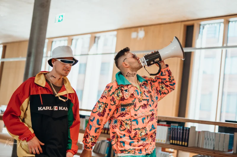 RawmanPictures - Sebastian Schiefermayr | Musikvideodreh "Flinz Asozial" | Zwei Männer im bunten Streetwear-Look mit Megafon in moderner Bibliotheksszene.
