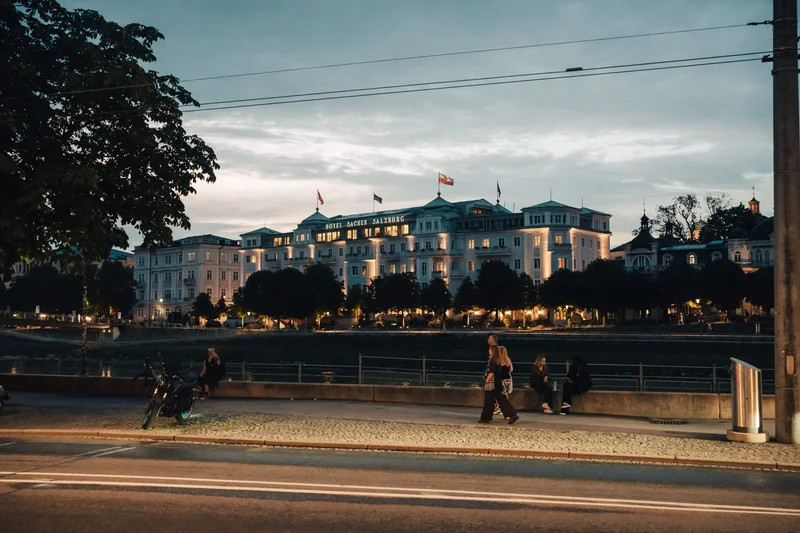RawmanPictures - Sebastian Schiefermayr | Contentday Salzburg | Abendliche Stadtansicht mit beleuchtetem Hotelgebäude und einer Flusslandschaft im Vordergrund.