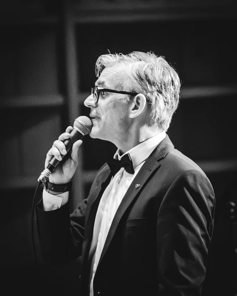 RawmanPictures - Sebastian Schiefermayr | Maturaball HTL1 | Schwarzweiß-Porträt eines Sängers auf der Bühne, Mikrofon, Anzug, emotionale Auftrittsfotografie.