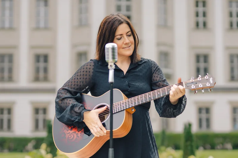 RawmanPictures - Sebastian Schiefermayr | Portraitshooting Tamara R. | Frau spielt Gitarre vor Gebäude, Musikfotografie, Open-Air-Konzert, Musikerin Outdoor-Auftritt.