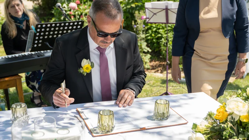RawmanPictures - Sebastian Schiefermayr | Hochzeit Traude & Gerhard | Mann im Anzug unterschreibt Heiratsurkunde bei einer Hochzeit im Freien.
