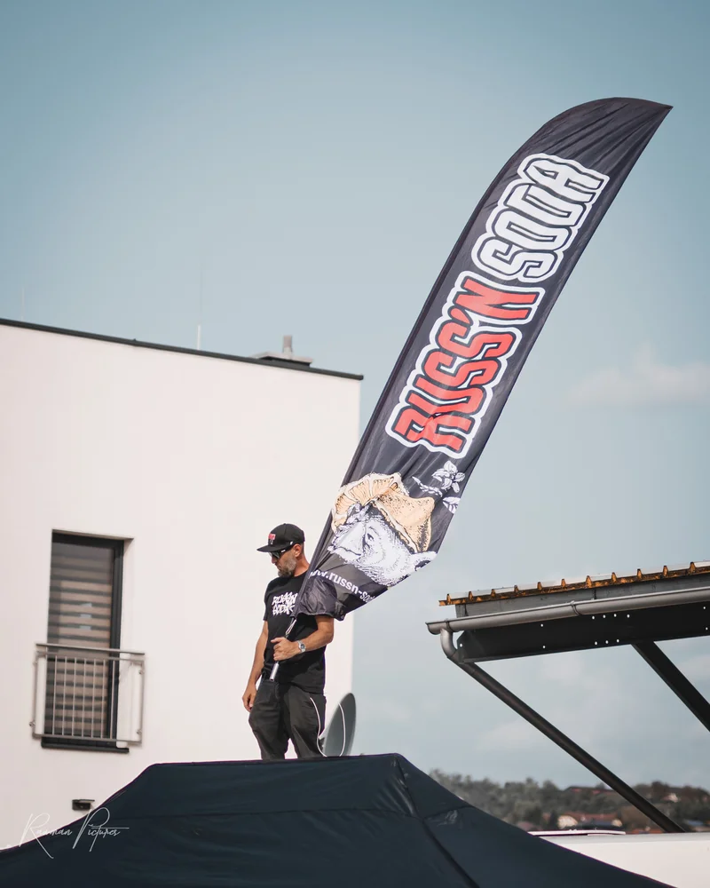 RawmanPictures - Sebastian Schiefermayr | Russnsoda Summer Closing | Outdoor-Eventfotografie: Mensch mit Flagge auf Dach in urbaner Kulisse, geeignet für Werbezwecke.