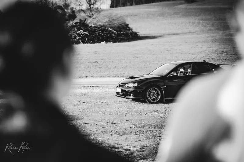 RawmanPictures - Sebastian Schiefermayr | Carshooting Eidenberg | Schwarzer Sportwagen auf ländlicher Straße im Vordergrund verschwommener Personen.