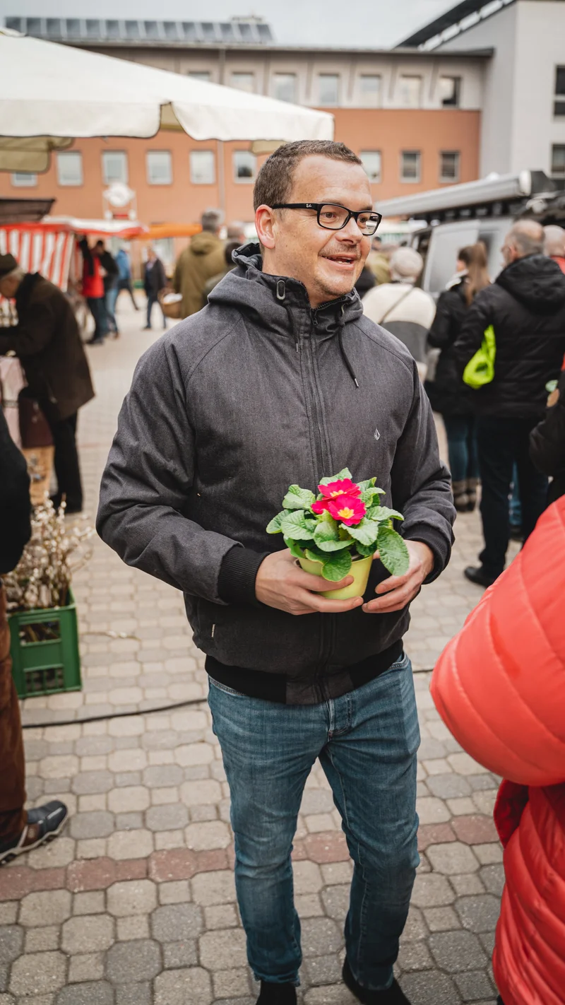 RawmanPictures - Sebastian Schiefermayr | Valentinsaktion Marchtrenk | Mann auf Markt mit Blume im Topf, lebendiger Outdoor-Fotografie-Moment.