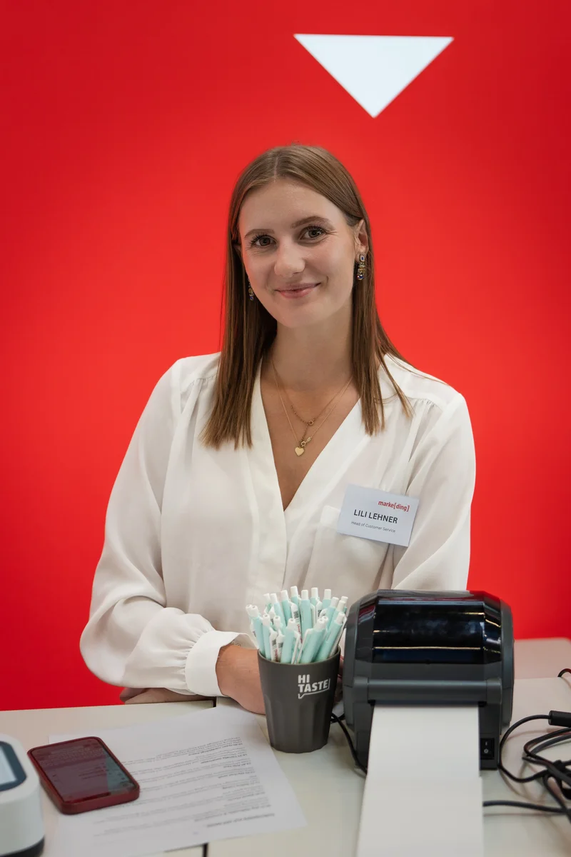 RawmanPictures - Sebastian Schiefermayr | Marke[ding] Wels | Messe-Standpersonal mit Lächeln vor rotem Hintergrund. Event-Fotografie mit professionellem Stil.