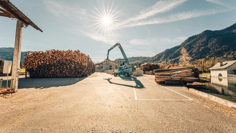 RawmanPictures - Sebastian Schiefermayr | Sägewerk Löberbauer | Sägewerk-Fotografie: Moderne Maschinen und Stapel von Holzstämmen in idyllischer Berglandschaft.