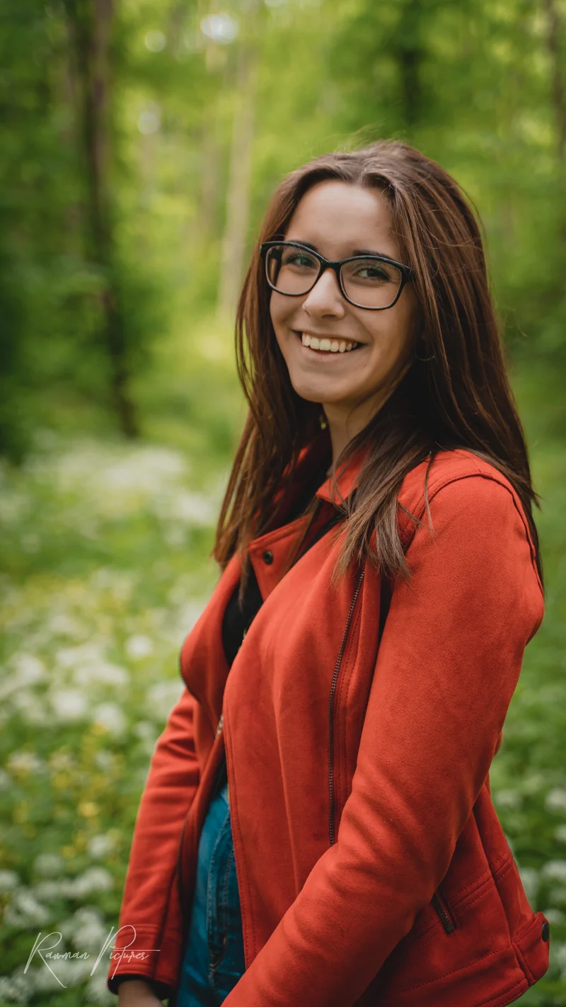 RawmanPictures - Sebastian Schiefermayr | Shooting Sophie N. | Porträtfotografie im Grünen: Frau mit Brille und roter Jacke lächelt im Wald.