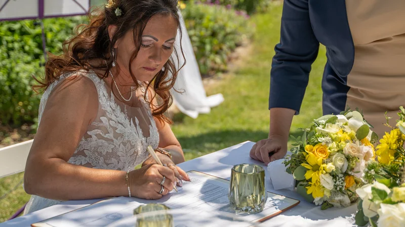 RawmanPictures - Sebastian Schiefermayr | Hochzeit Traude & Gerhard | Braut unterzeichnet Hochzeitsdokument im Freien, umgeben von Blumenarrangements.