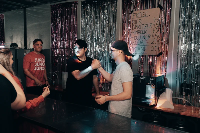 RawmanPictures - Sebastian Schiefermayr | JKU Mensafest | Menschen feiern an einer Bar mit Getränkepreisen im Hintergrund, lebendige Nachtlebenfotografie.