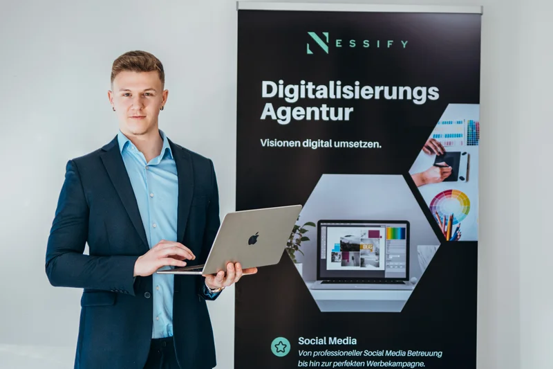 RawmanPictures - Sebastian Schiefermayr | Bussines-Shooting Nessify | Junger Unternehmer mit Laptop vor Banner einer Digitalisierungsagentur. Business-Portraitfotografie.