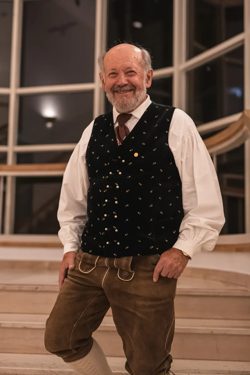 RawmanPictures - Sebastian Schiefermayr | Volkstanzfest | Portrait eines lächelnden Mannes in traditioneller Tracht vor eleganter Kulisse.