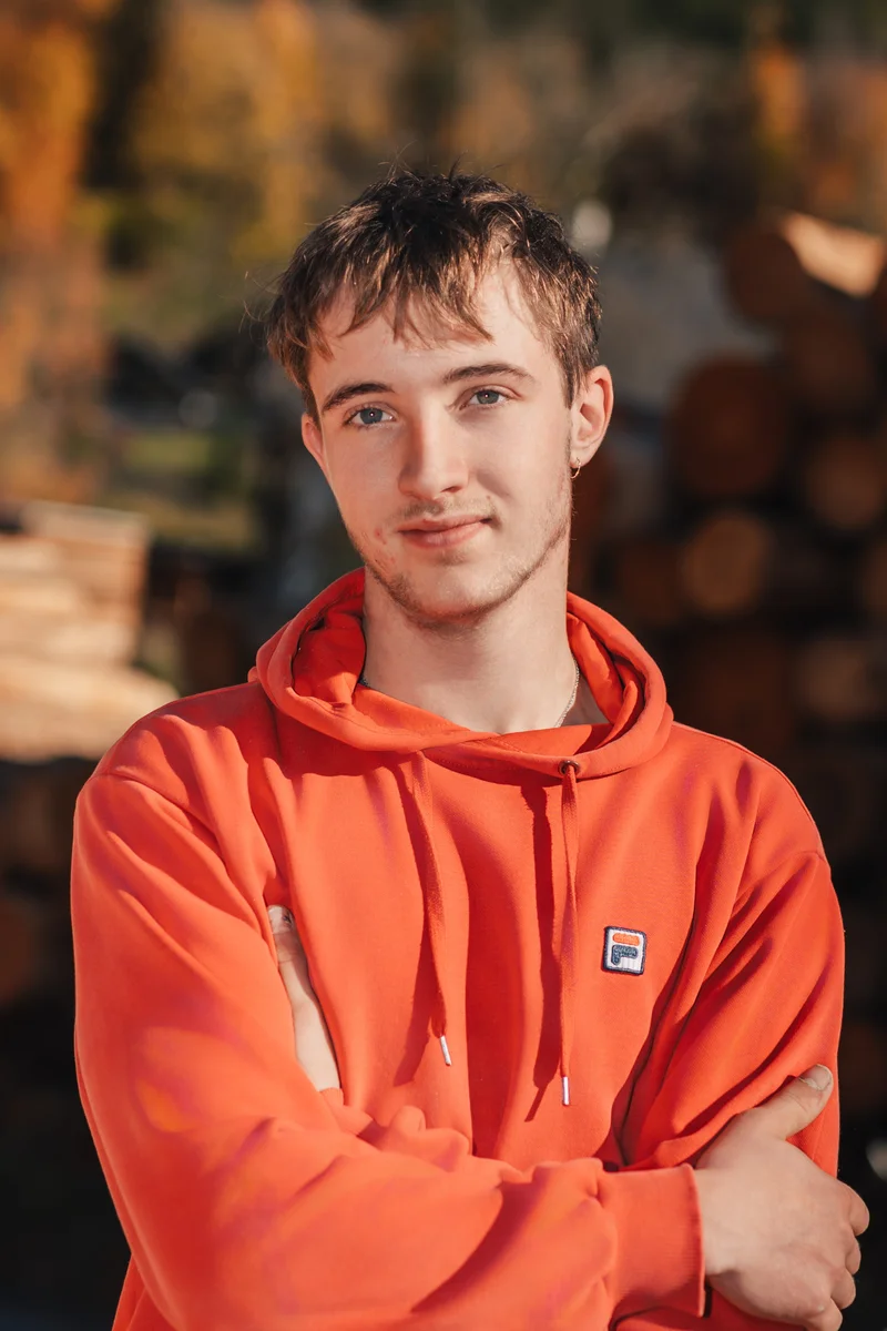 RawmanPictures - Sebastian Schiefermayr | Sägewerk Löberbauer | Junger Mann in rotem Hoodie lächelt draußen im Herbstlicht. Porträtfotografie im Freien.