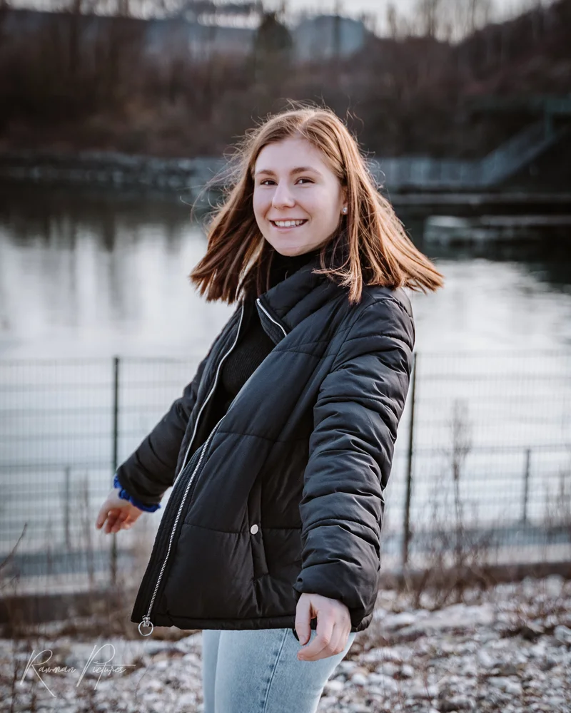 RawmanPictures - Sebastian Schiefermayr | Shooting Lea P. | Junge Frau lächelt am Flussufer, Wintermode, Outdoor-Porträtfotografie.