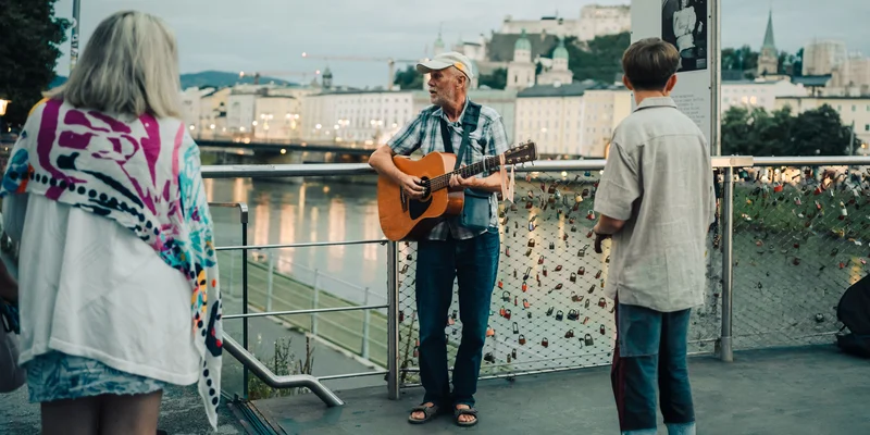 RawmanPictures - Sebastian Schiefermayr | Contentday Salzburg | Straßenmusiker mit Gitarre an einem Fluss; urbane Kulturfotografie in malerischer Abendstimmung.