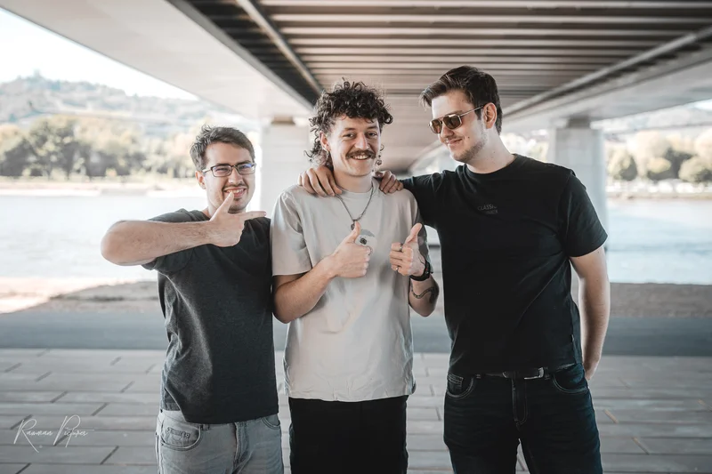 RawmanPictures - Sebastian Schiefermayr | Shooting Alex, Markus & Max | Drei Männer posieren lächelnd unter einer Brücke am Flussufer. Stadtfotografie, Freundschaft.