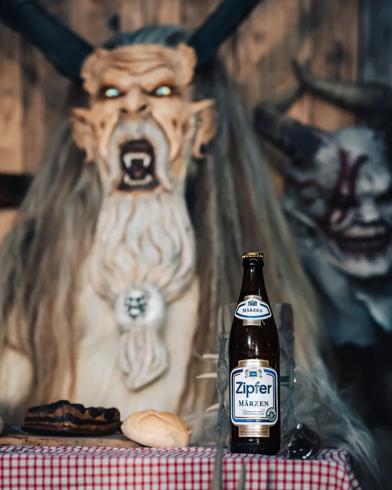 RawmanPictures - Sebastian Schiefermayr | Maskenausstellung | Gruselige Krampus-Dekoration mit Zipfer Märzen Bierflasche auf traditionellem Tischdisplay.