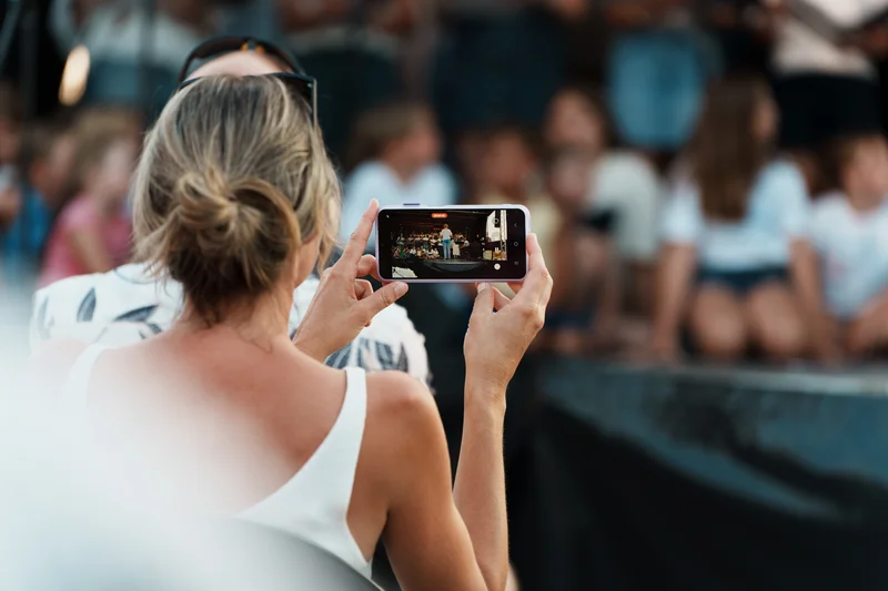 RawmanPictures - Sebastian Schiefermayr | Singkreis-Sundowner  | Eventfotografie: Frau filmt Konzert mit Smartphone, erfasst lebendige Bühnenatmosphäre.