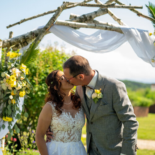 RawmanPictures - Sebastian Schiefermayr | Hochzeit Traude & Gerhard | Romantische Hochzeitsfotografie im Freien, Brautpaar unter Blumenbogen, strahlender Sonnenschein.
