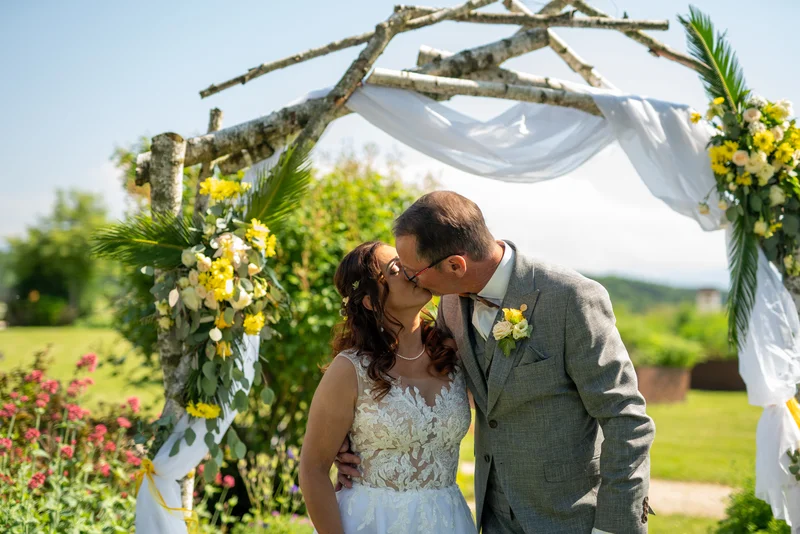 RawmanPictures - Sebastian Schiefermayr | Hochzeit Traude & Gerhard | Romantische Hochzeitsfotografie im Freien, Brautpaar unter Blumenbogen, strahlender Sonnenschein.