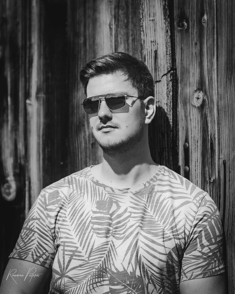 RawmanPictures - Sebastian Schiefermayr | Kirschblüten­wanderung  | Junger Mann in Sonnenbrille, Portraitfotografie vor Holzwand, Schwarz-Weiß.