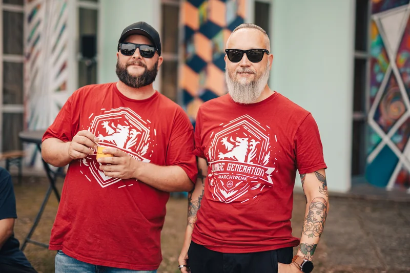 RawmanPictures - Sebastian Schiefermayr | Sommereisdisco | Zwei Männer bei einem Event in rot bedruckten T-Shirts mit Sonnenbrillen und Bart, fröhlich posierend.