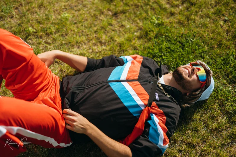 RawmanPictures - Sebastian Schiefermayr | Musikvideodreh "Flinz Asozial" | Hipster im Vintage-Sportoutfit liegt entspannt auf grüner Wiese, sonnige Outdoor-Fotografie.