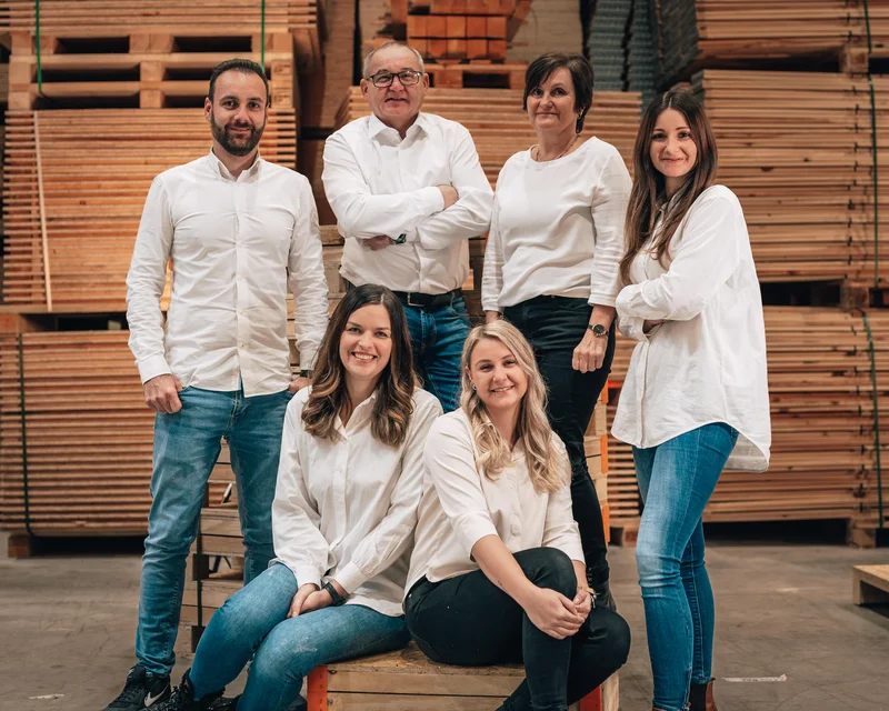 RawmanPictures - Sebastian Schiefermayr | Firmenshooting EasyBox GmbH | Gruppenfoto in Holzwerkstatt: Team im weißen Outfit vor gestapeltem Holz für Firmenwebsite.