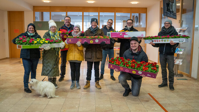 RawmanPictures - Sebastian Schiefermayr | Valentinsaktion Marchtrenk | Gruppenfoto mit Blumen: Innenaufnahme, fröhliche Menschen, Primeln, Winterkleidung, Gemeinschaft.