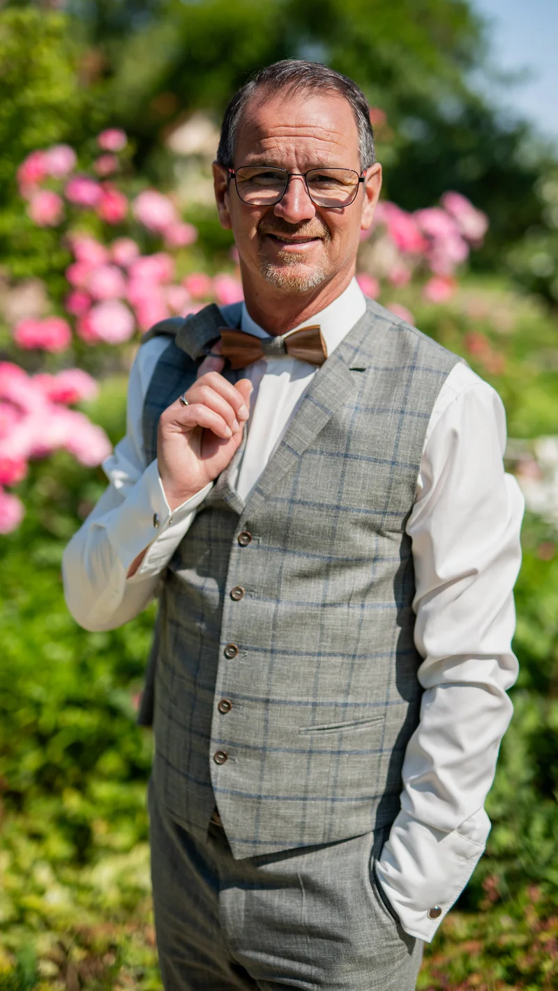 RawmanPictures - Sebastian Schiefermayr | Hochzeit Traude & Gerhard | Eleganter Herr im Anzug lächelt im Garten. Herrenporträt Fotografie, Outdoor-Setting.