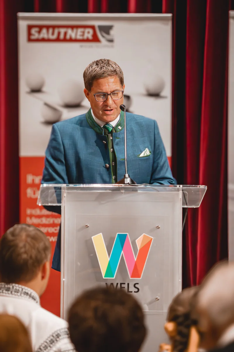 RawmanPictures - Sebastian Schiefermayr | Volkstanzfest | Redner bei einer Veranstaltung: Sprechender Mann am Podium mit Mikrofon vor Publikum.