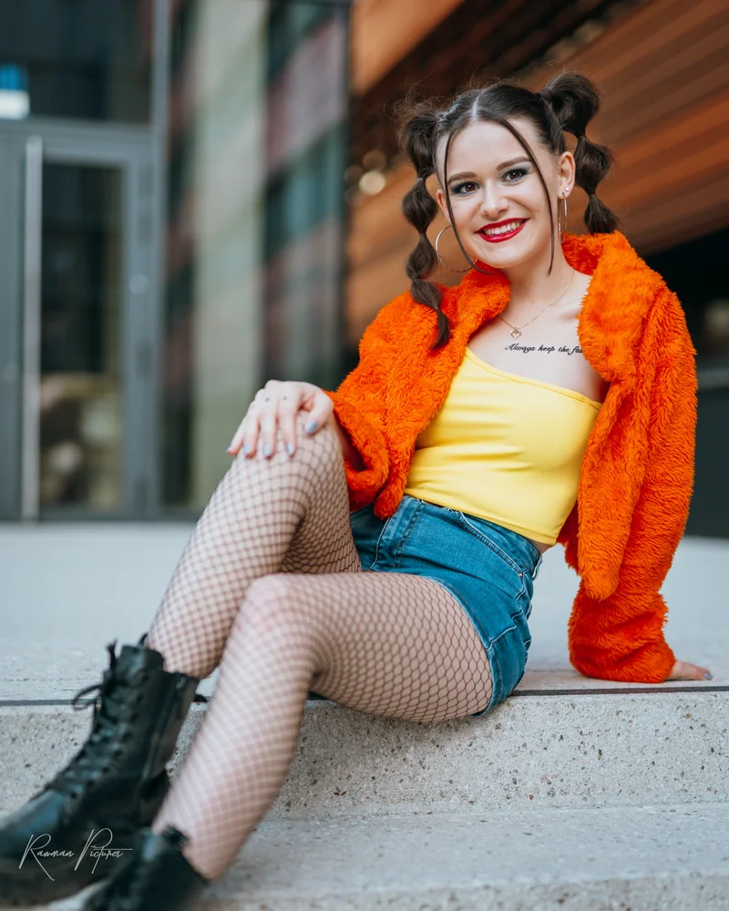 RawmanPictures - Sebastian Schiefermayr | Musikvideodreh "Flinz Asozial" | Junge Frau in urbanem Outfit, fröhlich draußen sitzend, moderne Portraitfotografie.