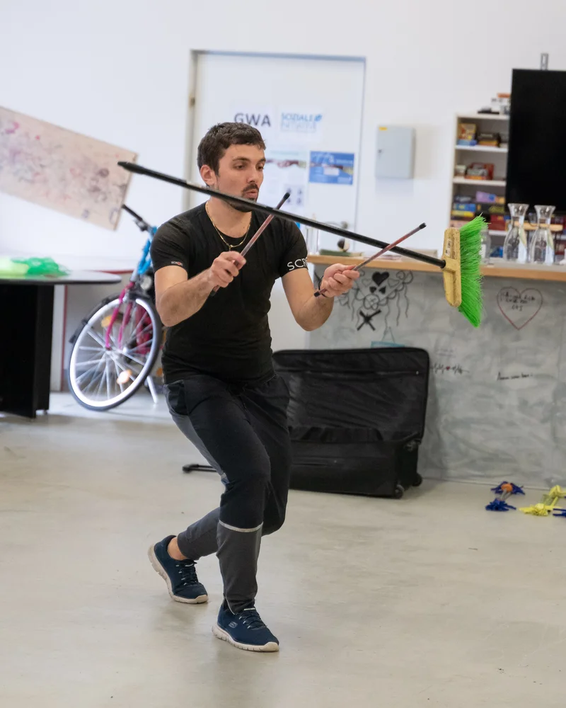 RawmanPictures - Sebastian Schiefermayr | Jonglierworkshop Marchtrenk | Mann beim Jonglieren mit Besen und Besenstiel in einem Raum voller Requisiten.