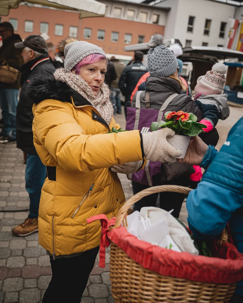 RawmanPictures - Sebastian Schiefermayr | Valentinsaktion Marchtrenk | Frau auf Markt kauft bunte Blumen, winterliche Szene, Einkaufsatmosphäre.