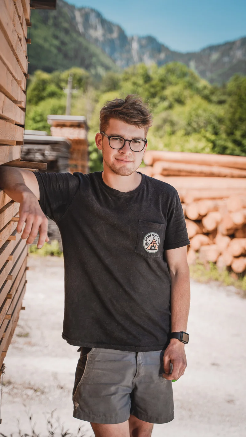 RawmanPictures - Sebastian Schiefermayr | Sägewerk Löberbauer | Person outdoors mit Holzstapeln und Bergen im Hintergrund. Perfekt für Portraitfotografie.