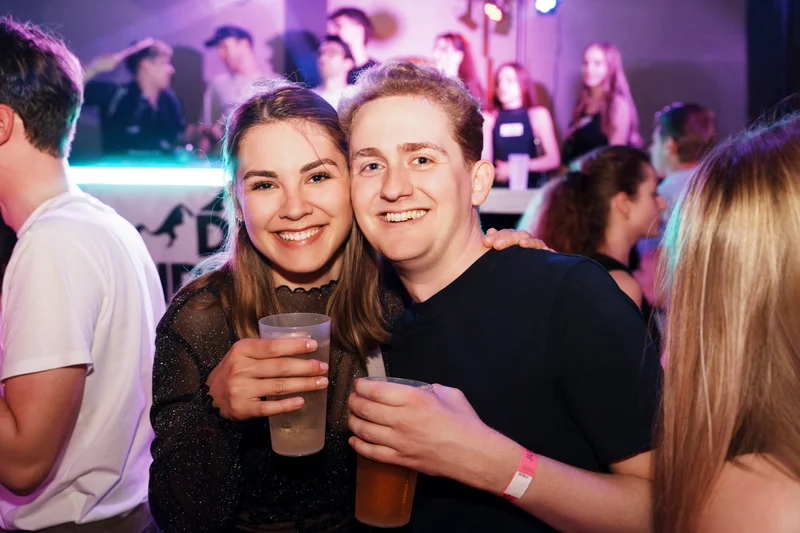 RawmanPictures - Sebastian Schiefermayr | JKU Mensafest | Fröhliche Partystimmung mit Freunden, lächelnd mit Getränken in lebhafter Clubatmosphäre.