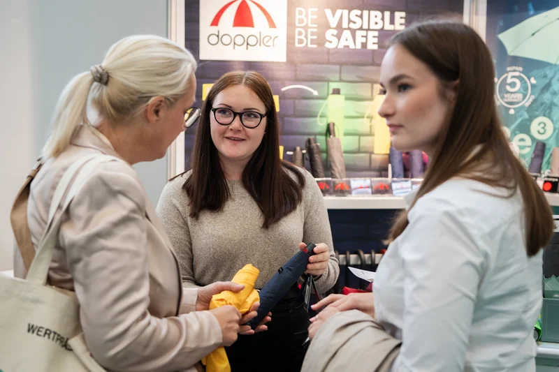 RawmanPictures - Sebastian Schiefermayr | Marke[ding] Wels | Drei Frauen im Gespräch auf einer Messe über sichere und sichtbare Regenschirme.