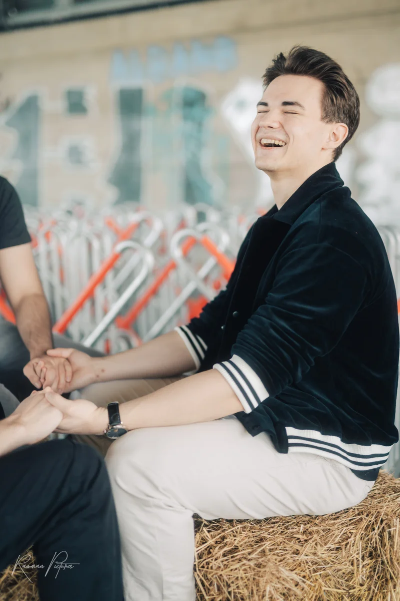 RawmanPictures - Sebastian Schiefermayr | Shooting Alex, Markus & Max | Lachender Mann sitzt auf Heuballen, urbane Kulisse, authentische Momente, Lifestyle-Fotografie.