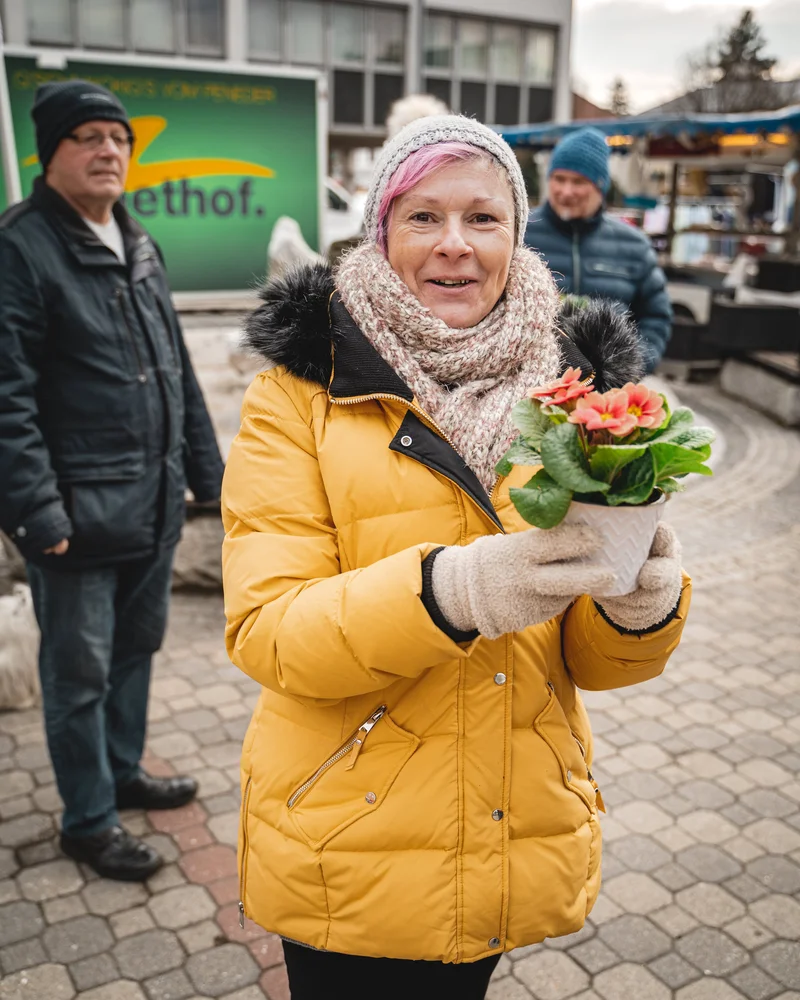 RawmanPictures - Sebastian Schiefermayr | Valentinsaktion Marchtrenk | Eine Frau in Winterkleidung hält eine Blume auf einem belebten Marktplatz.